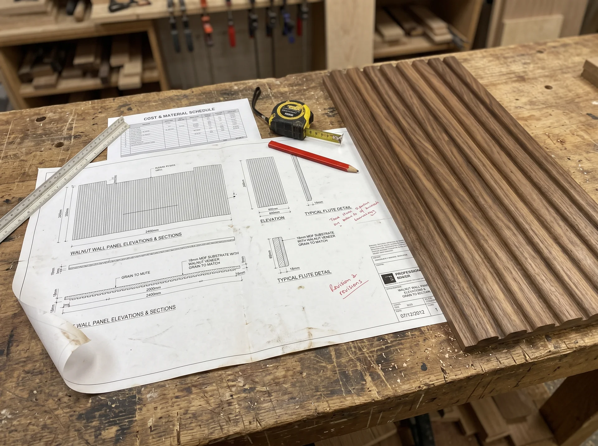 A joinery shop drawing of walnut wall panels with its cost and material schedule, laid beside the finished fluted panel on a workbench